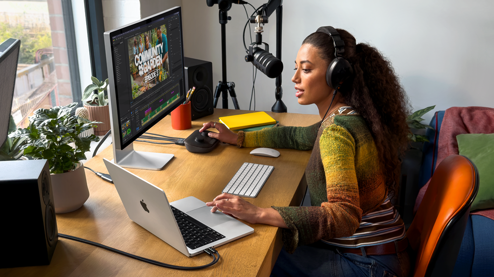 Una mujer en su casa trabaja en su mesa con un MacBook Pro conectado a un monitor externo, lleva auriculares y habla por un micrófono