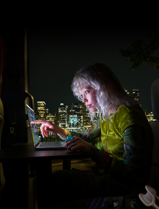 Una persona sentada en el asiento trasero de un furgón, la puerta del pasajero abierta muestra el horizonte de una ciudad por la noche, MacBook Pro con una pantalla PROLED XDR sobre una mesa de trabajo, la persona está iluminada por la pantalla mientras utiliza el control táctil para editar un vídeo en DaVinci Resolve Studio