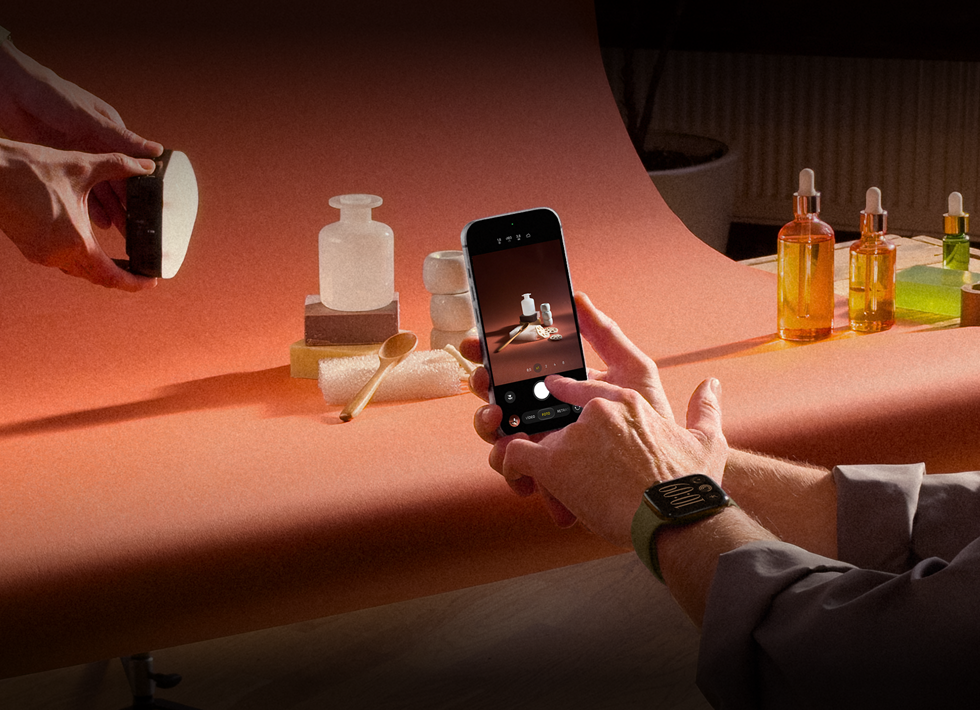 Sesión de fotos en un lugar de trabajo, una persona usa un iPhone para fotografiar productos sobre un fondo, otra sostiene una lámpara